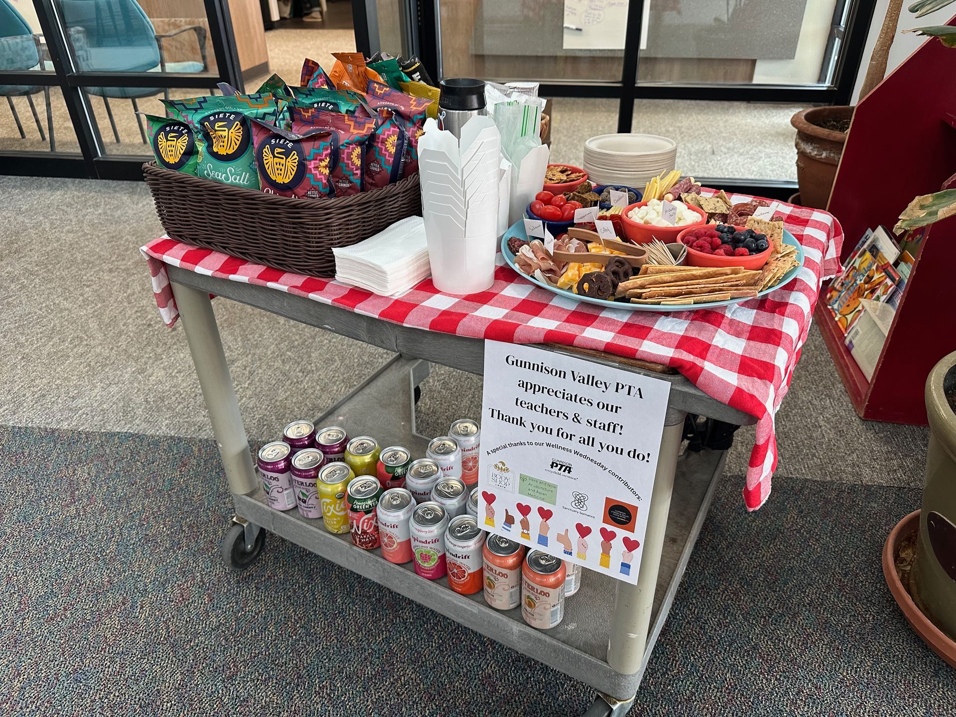 A snack cart with chips, charcuterie, and drinks. It has a red and white checkered tablecloth.