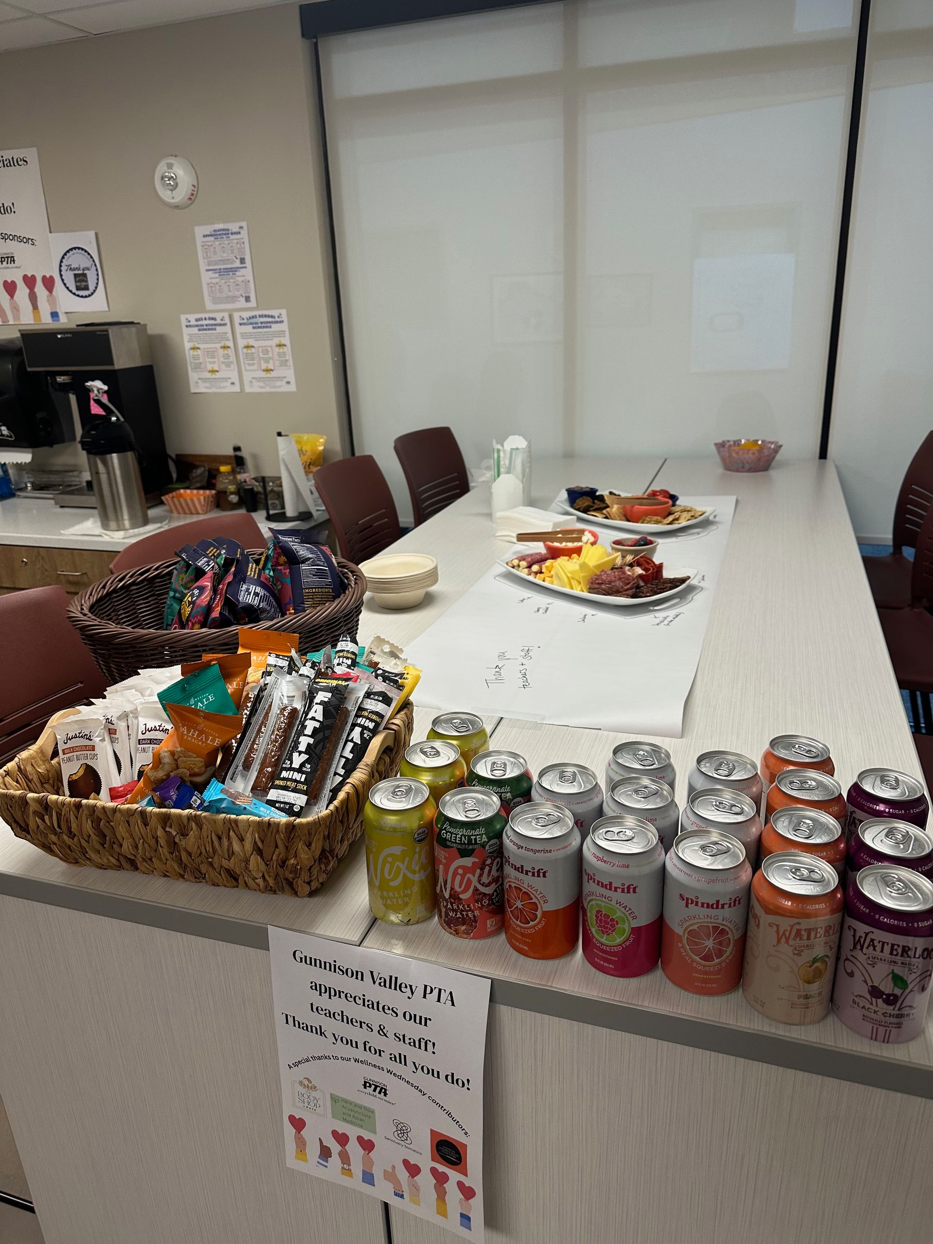 A table with snacks and drinks, including a basket of treats and cans of soda, in an office.