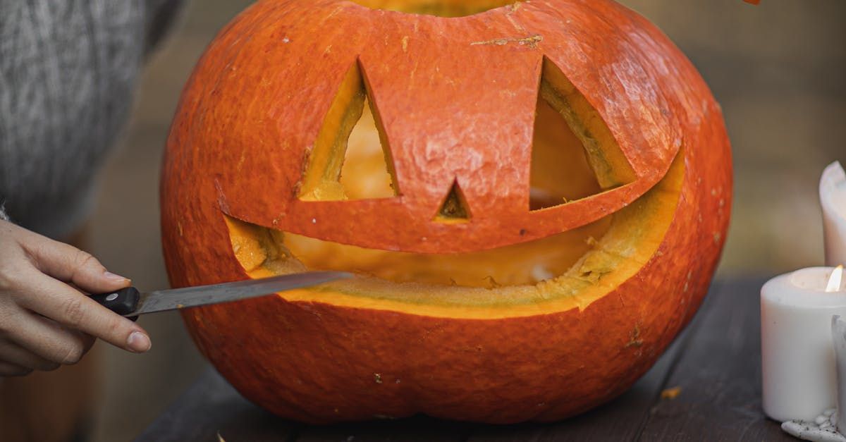 Person carving a jack-o'-lantern with a knife on a wooden surface, with lit candles in the background.