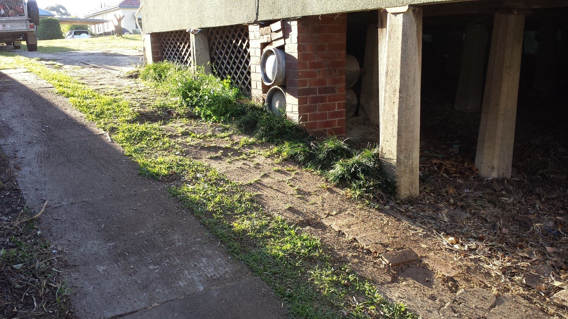 A side view of a house with visible foundation pillars and a walkway. The surrounding area includes grass and some weeds, indicating a need for maintenance. This image illustrates a property area potentially serviced by Abate Rubbish Removal's junk removal services in Newcastle.
