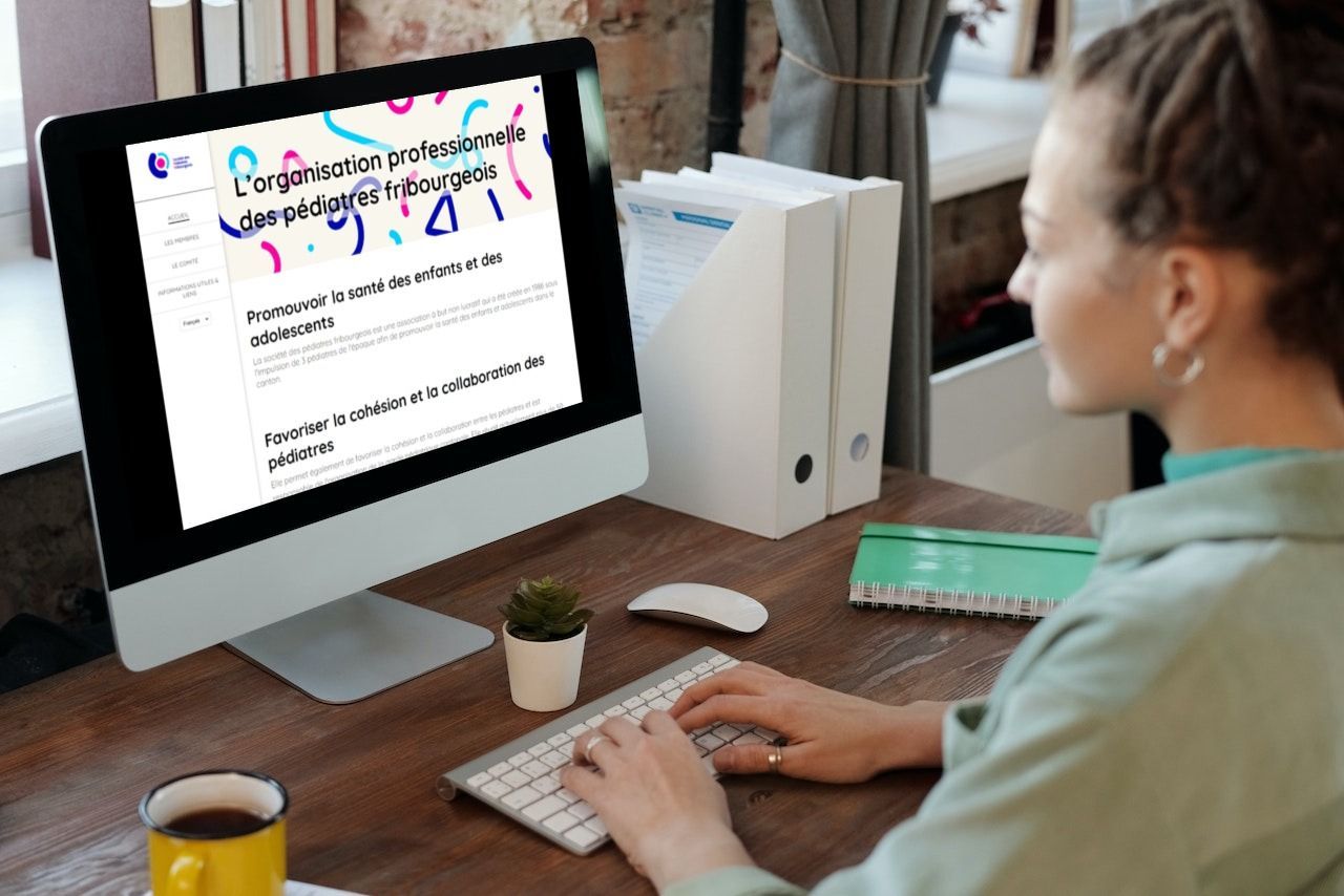 Une femme tape sur un clavier, consultant une page web sur un écran d'ordinateur, assise à son bureau avec un café et des dossiers.