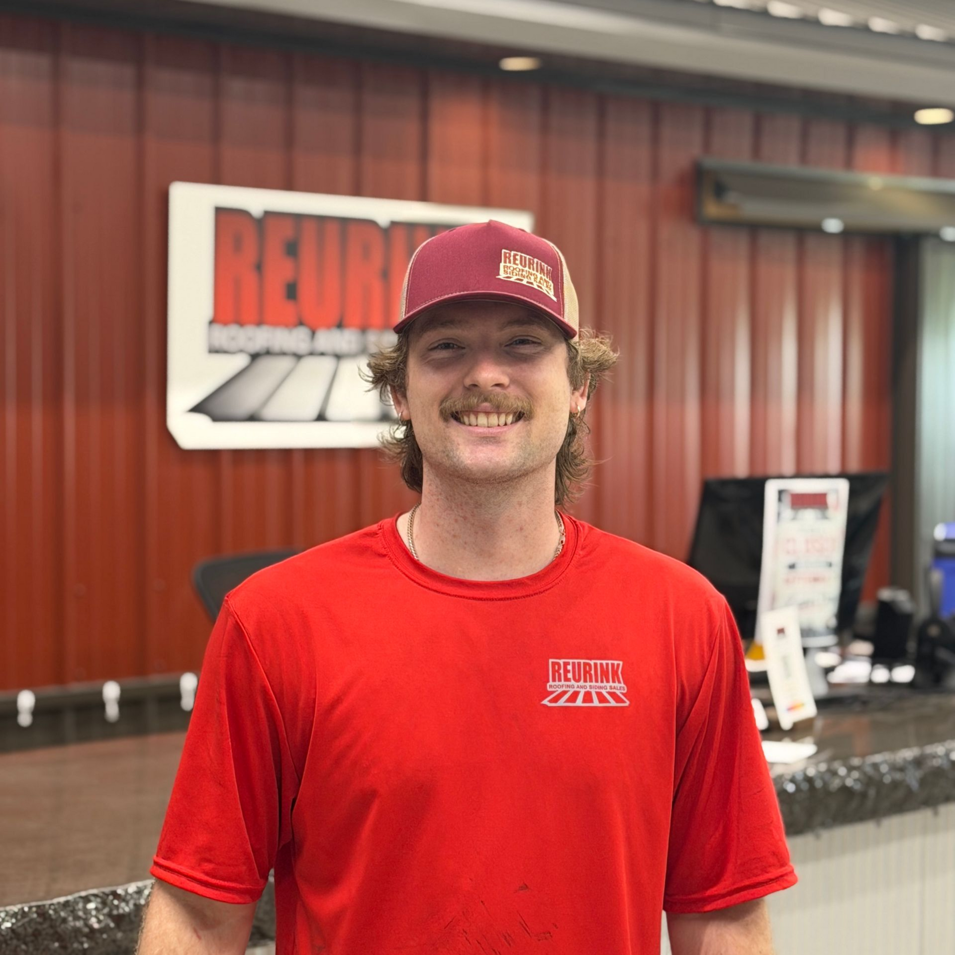 Man in red sweatshirt and green cap, smiling, with a radio mic. Standing in front of a building.