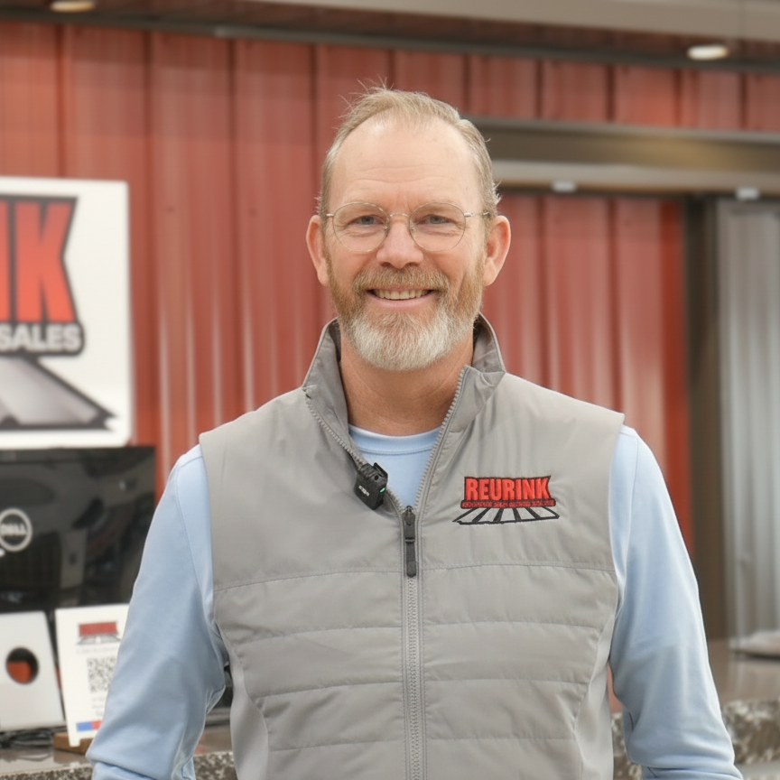 Man with glasses and beard wearing a gray vest in front of a sign that says
