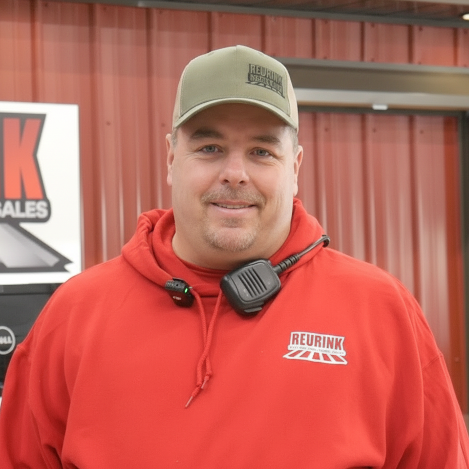 Man wearing red hoodie and green cap with a microphone. He stands in front of a red building.