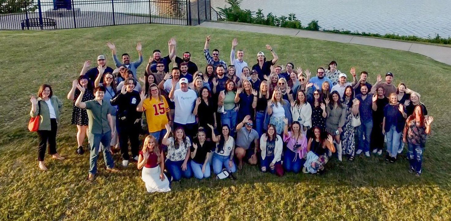 Large group of people on a grassy hill waving at the camera. A body of water and fence are in the background.