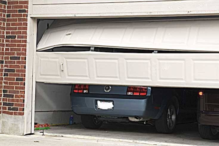 A car is stuck under a partially open, damaged garage door.