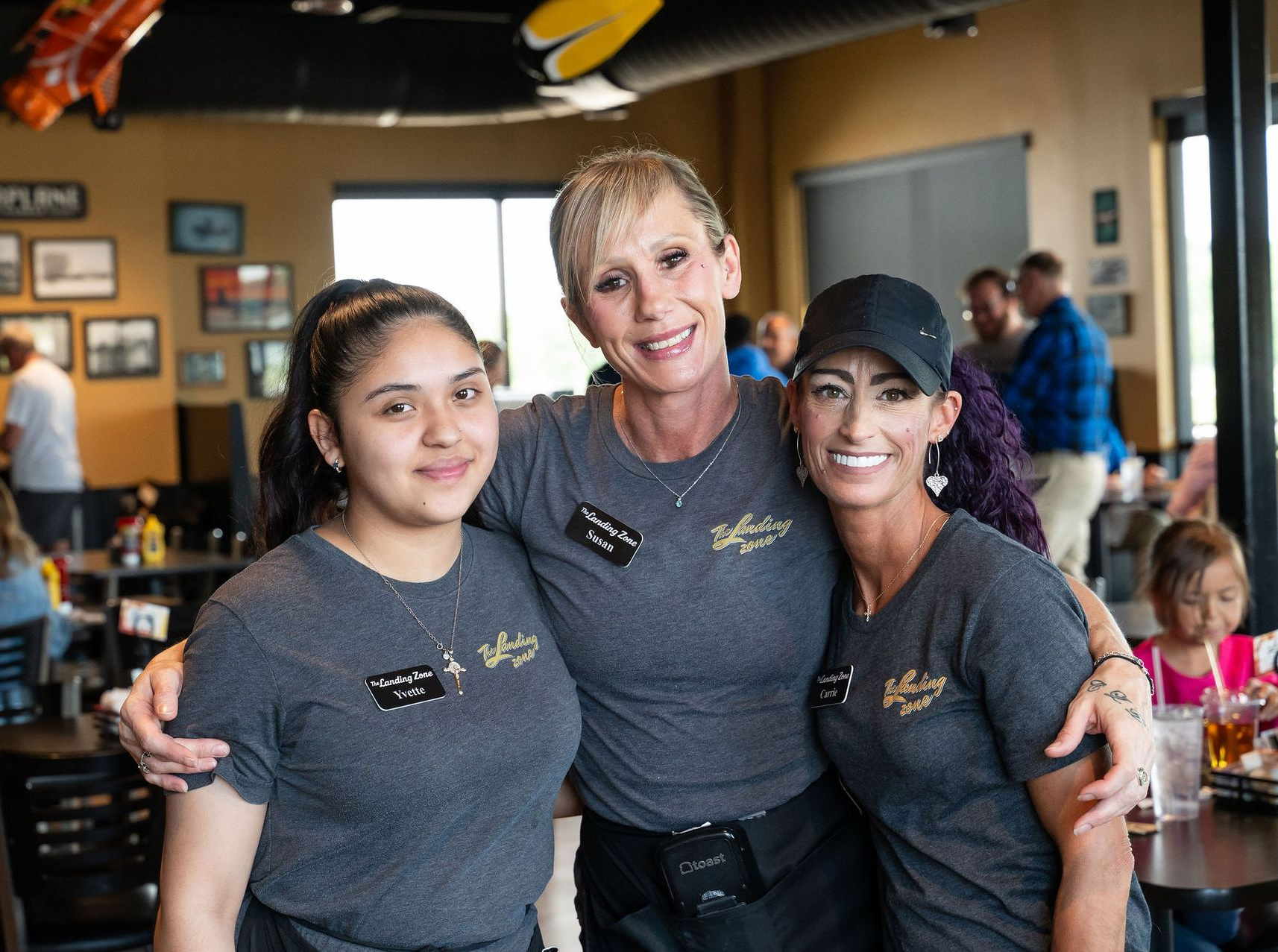 Three people in gray shirts smiling inside a lively restaurant or cafe.