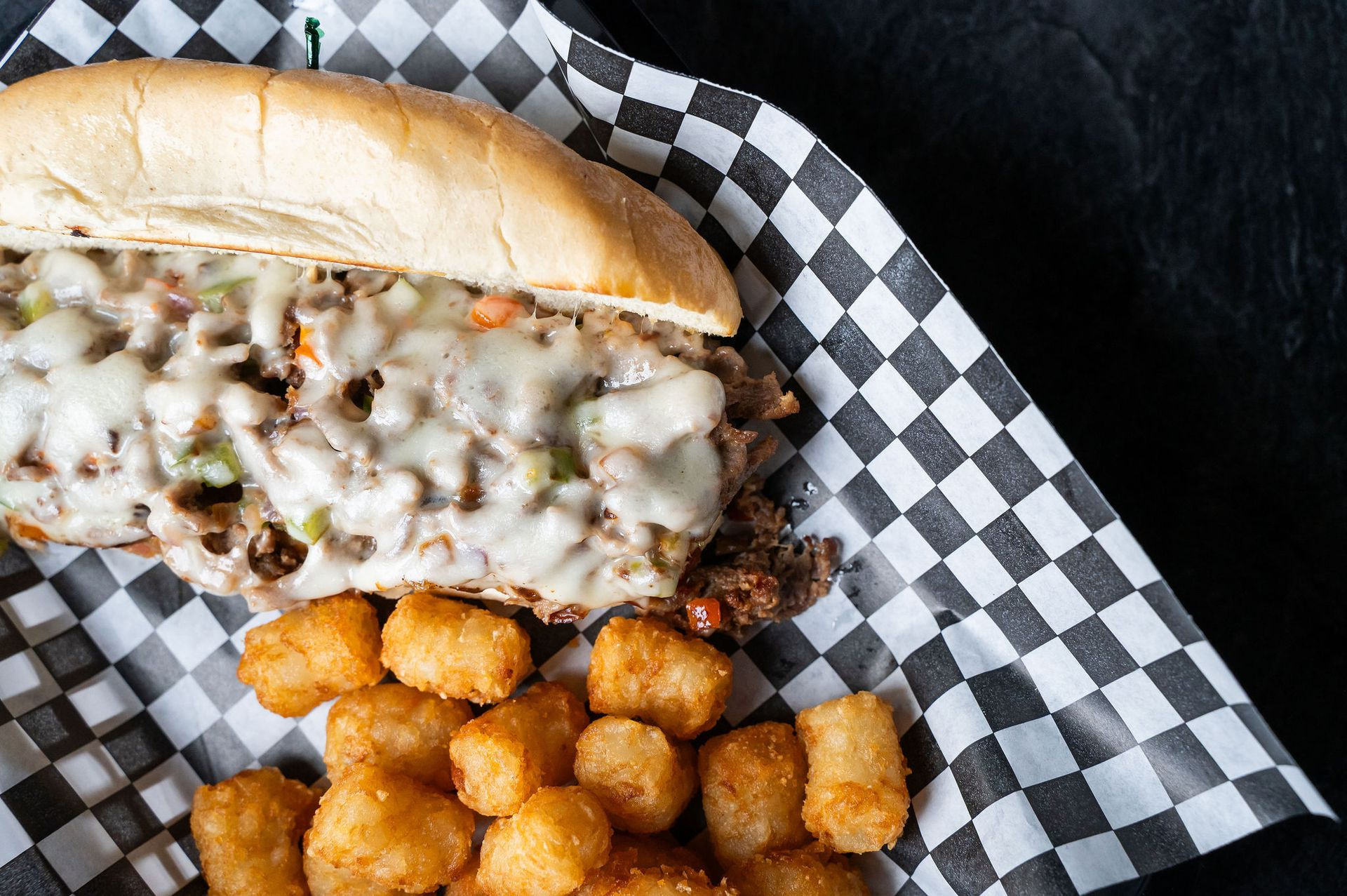 Cheesesteak sandwich with melted cheese and tater tots on checkered paper