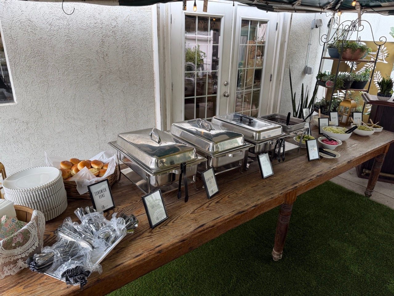 Outdoor buffet table with covered trays, bowls, and labeled dishes under a white canopy.