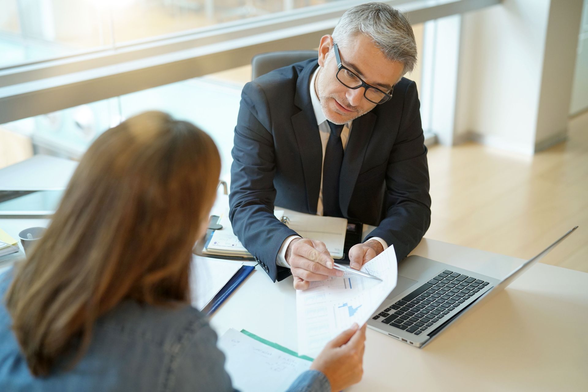 Two people in a bright office discuss a document across a table with a laptop open nearby.