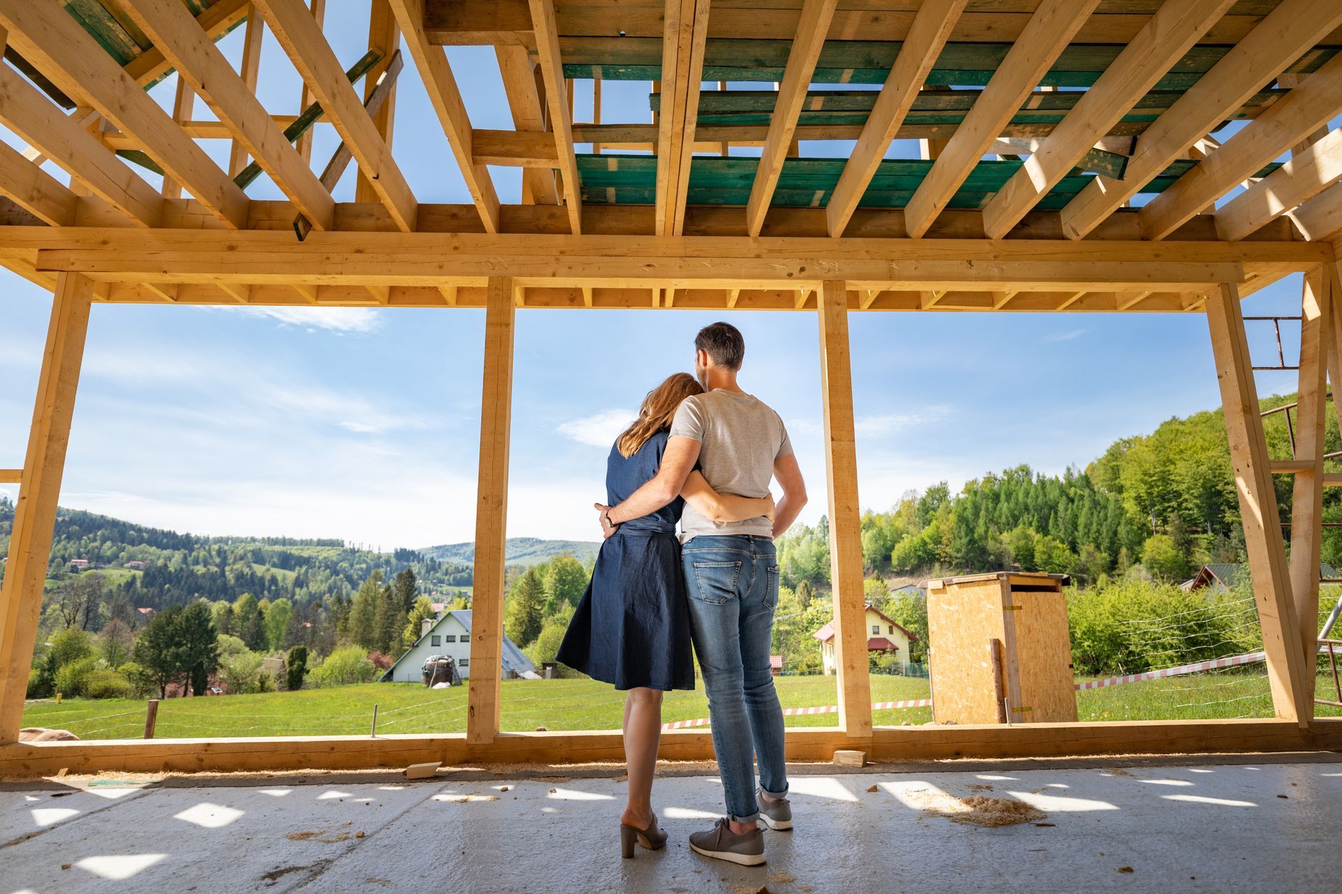Couple embracing inside a wooden-framed house under construction, with a green valley view outside.