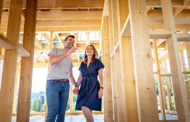 Two people standing in a framed wooden house under construction, looking at plans and smiling