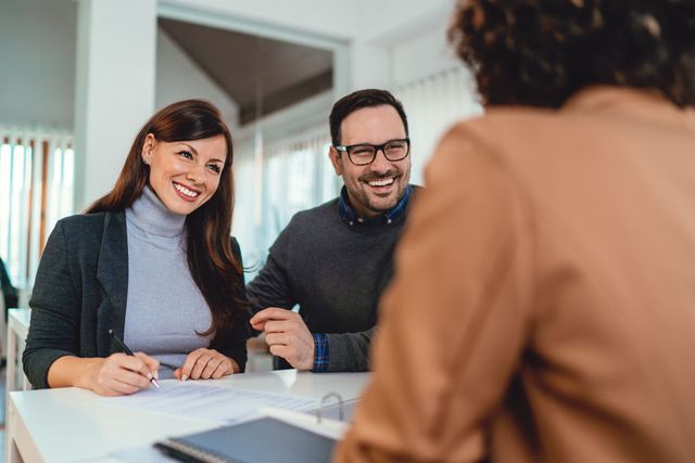 Two people smiling at a desk in a meeting with a person speaking in the foreground