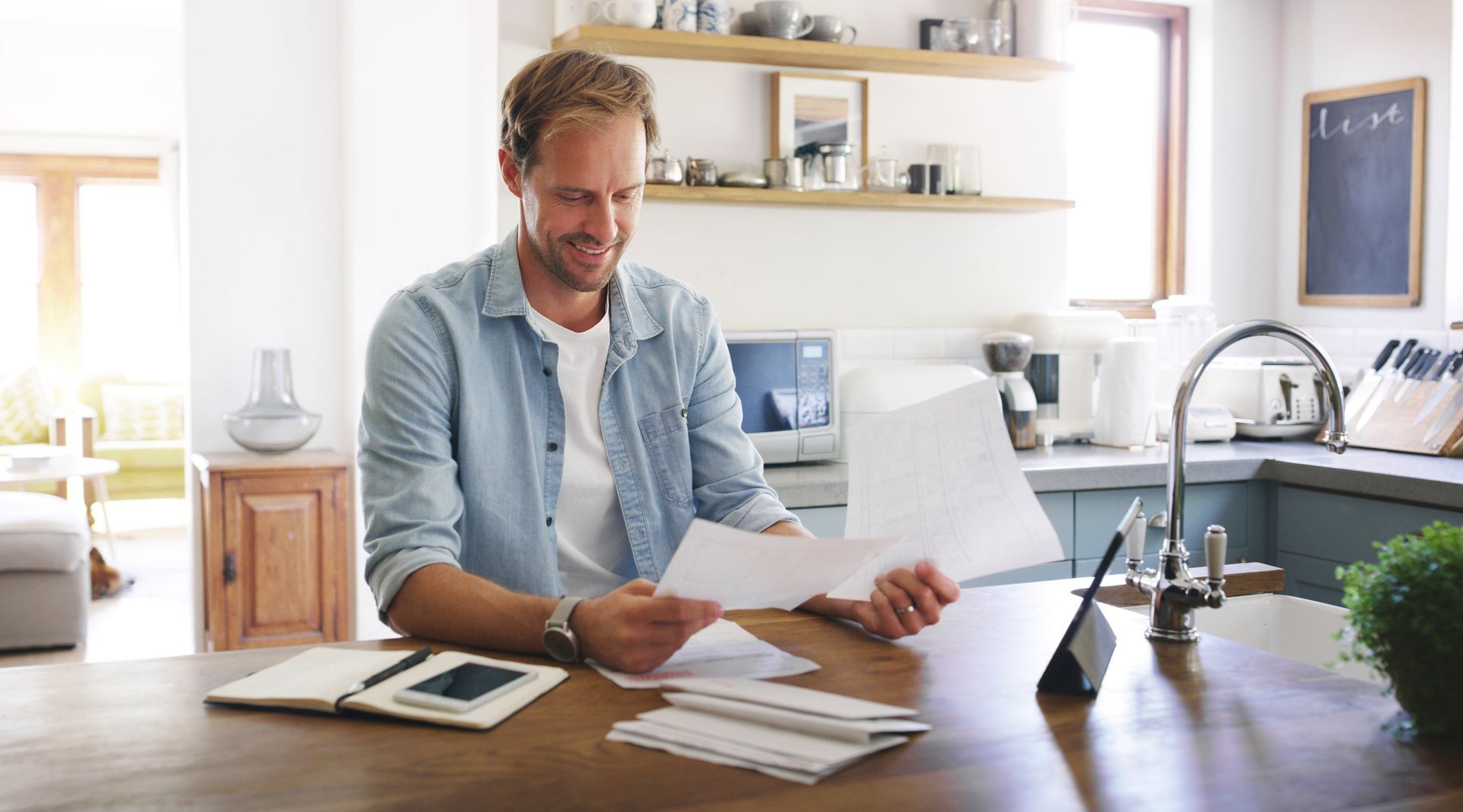 Man sitting at a kitchen table sorting papers, with a tablet, coffee, and shelves in the bright background