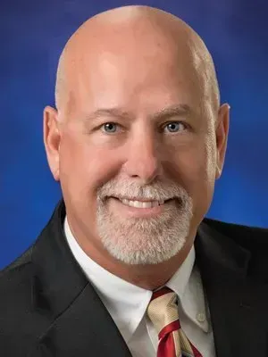 Smiling bald man in a dark suit and striped tie against a blue background