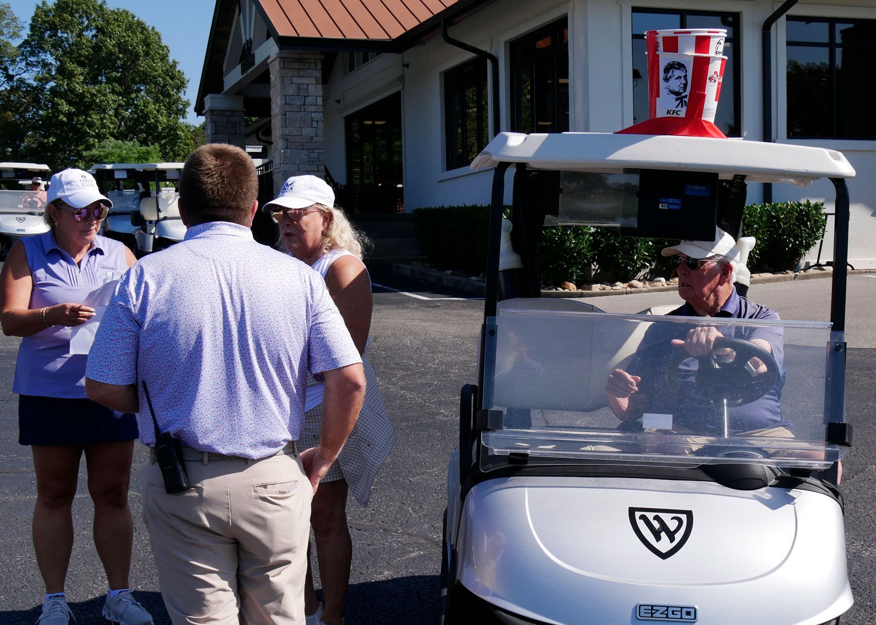 Debbie Baldwin, Josh Griffin, Diane Nelson, and Tom Briggs meeting before the scramble kicks off. Tom Briggs is sitting in his golf cart, adorned with a KFC bucket and the image of the Colonel has been replaced with a likeness of Tom.