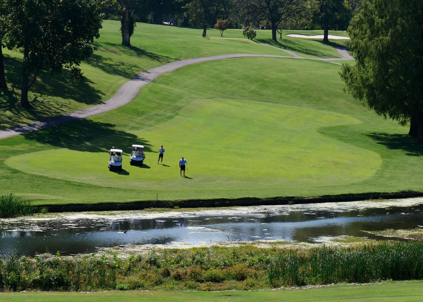 Two men and two golf carts on the fairway against a hill. In the foreground, stream runs through the fairway.