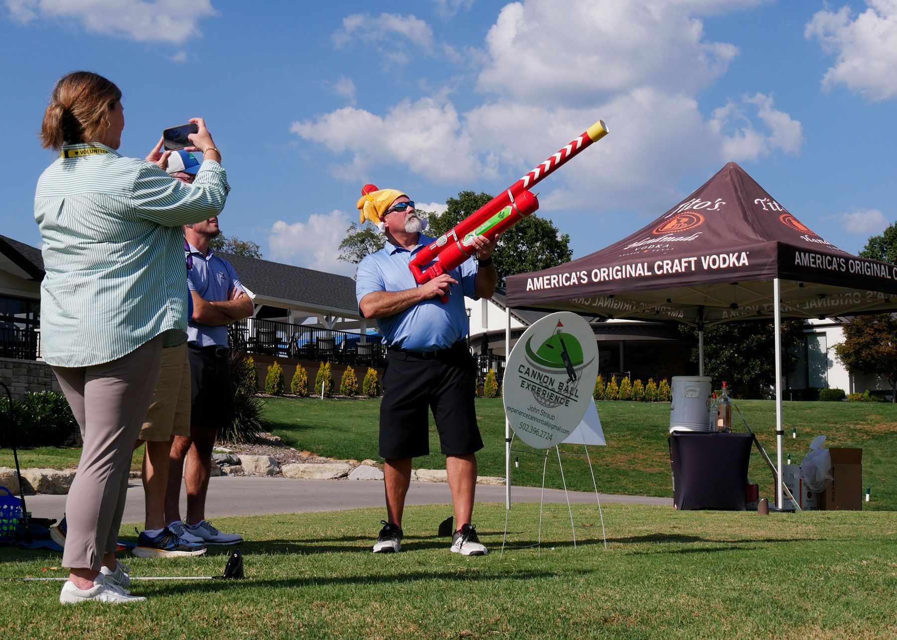 A man wearing a hat that looks like a roasted chicken shoots a golf ball from the air cannon.