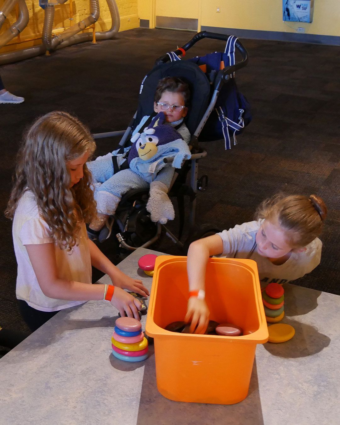 At one of the interactive displays at the Kentucky Science Center, two girls stack small discs of varying size and color while a small boy watches from his stroller.