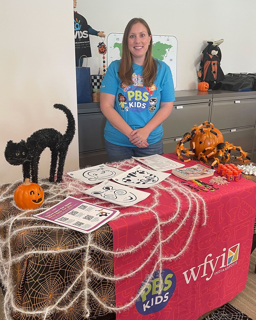 A woman representing WFYI/PBS Kids poses for a photo behind their table at the FRC, which is decorated with pumpkins, spiders, and a black cat.
