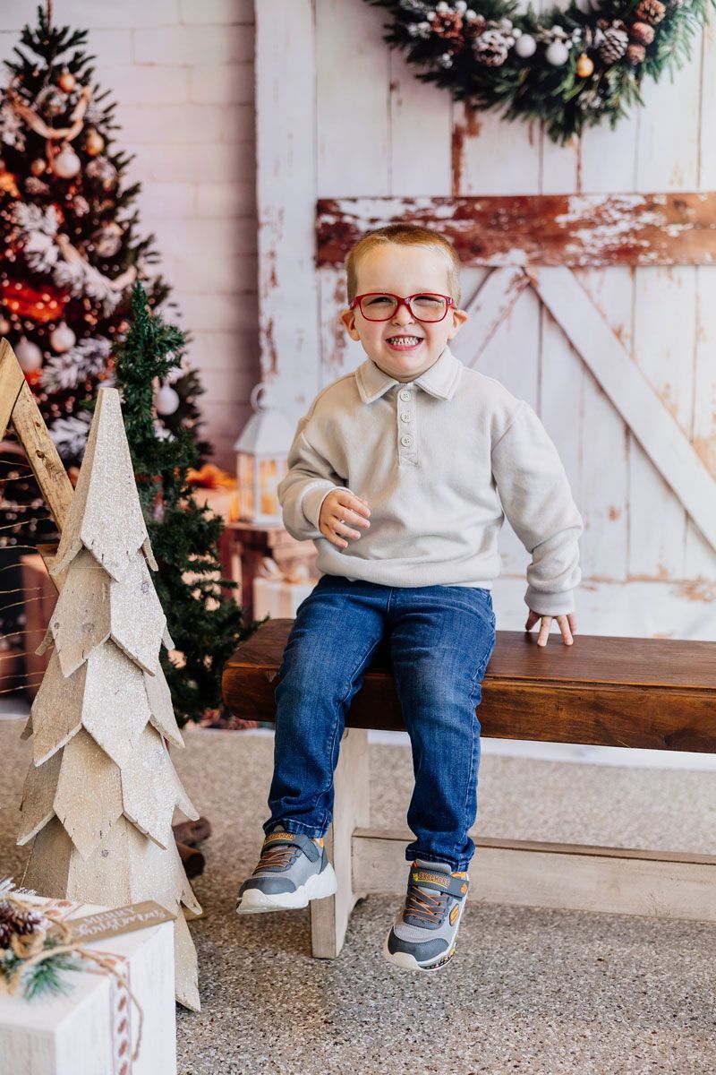 Clyde poses for a photo with a big smile and his new, red glasses, sitting on a wooden bench, with a Christmas tree in the background.