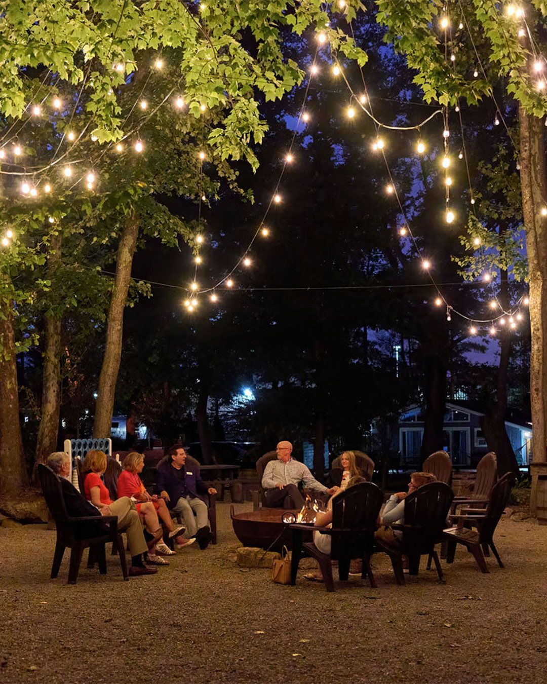 People sit around a fire pit under trees lit with string lights at Progress Park.