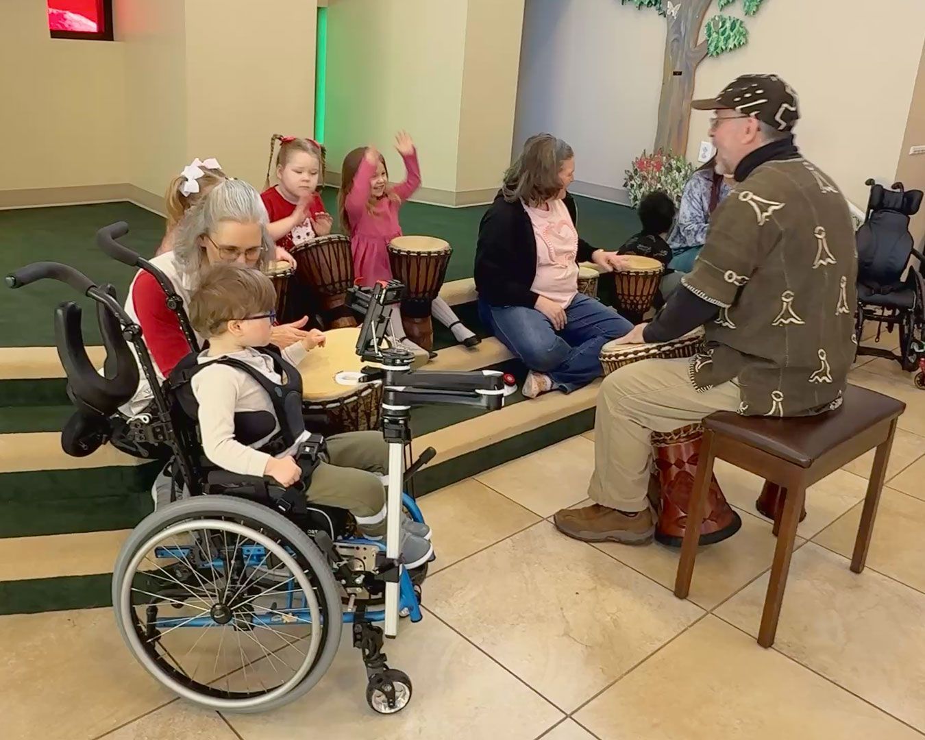 The kids sit on the steps in Kids Town and play drums with Gregory.