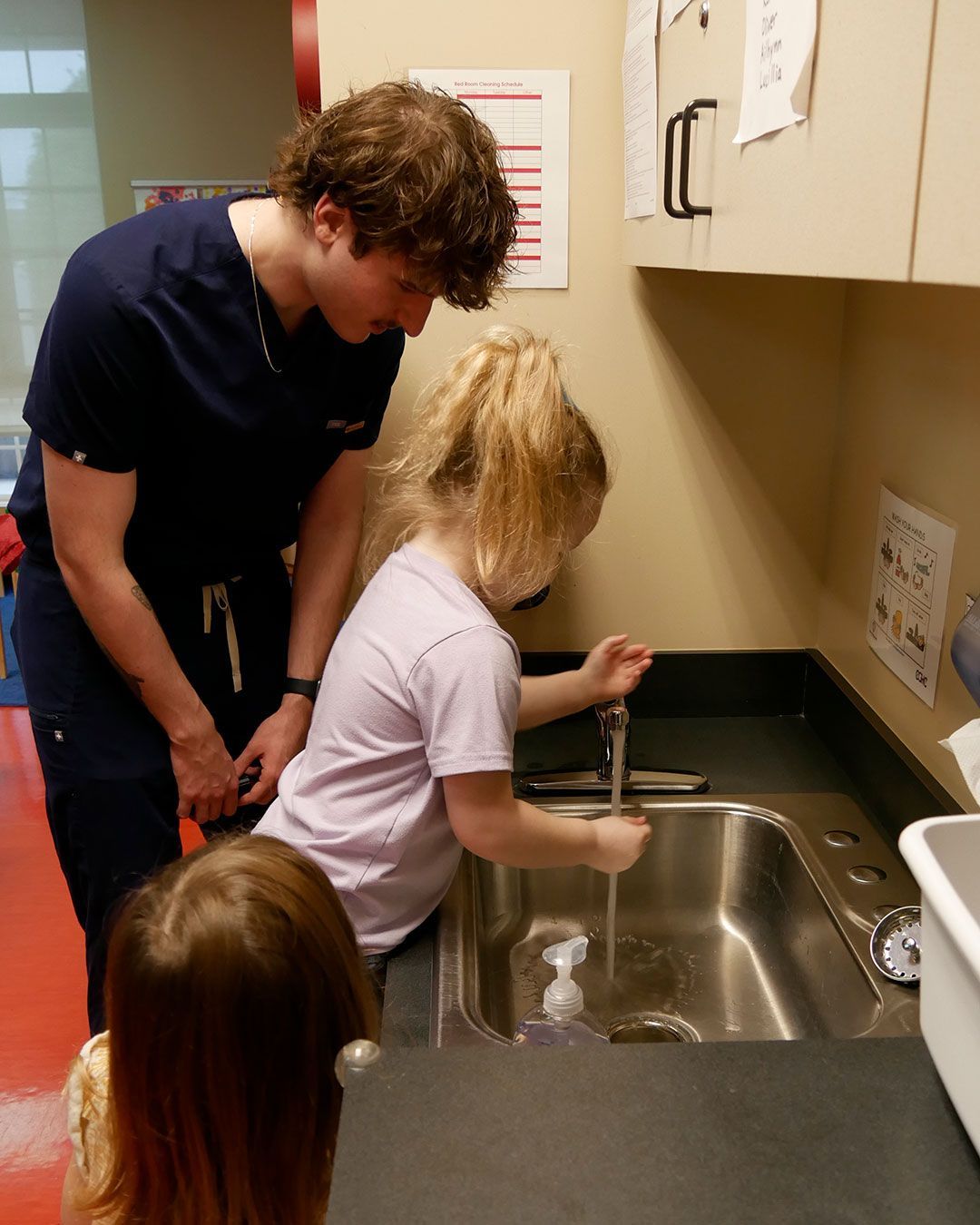 A small girl washes her hands under the guidance of a male physician assistant student.