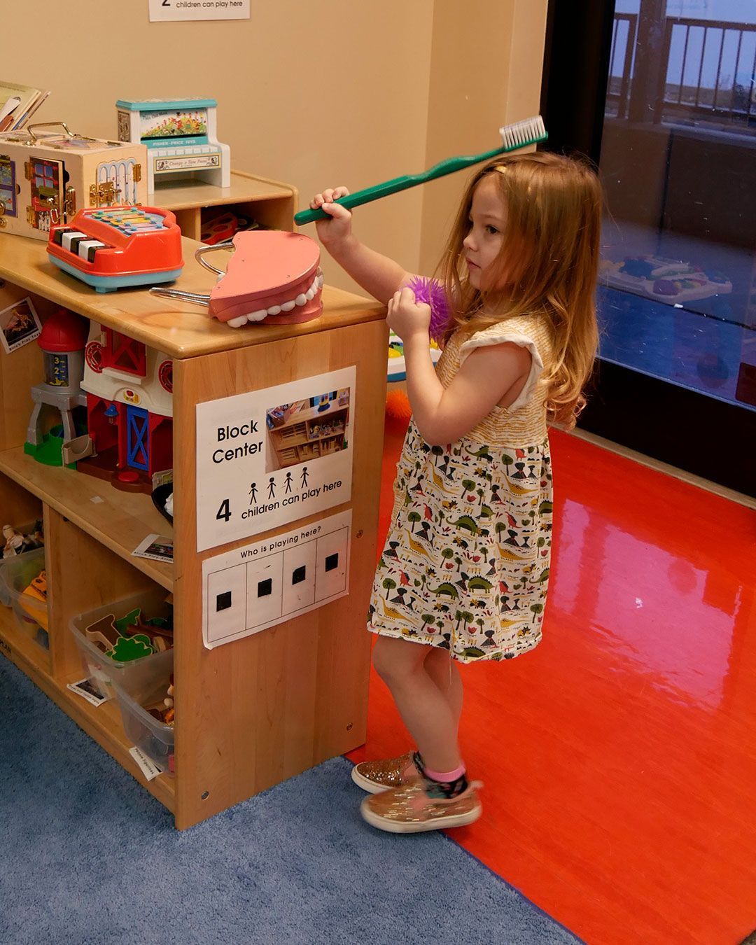 A small girl holds a giant toothbrush in one hand and her 'germ' toy in another while standing in front of a giant set of teeth.