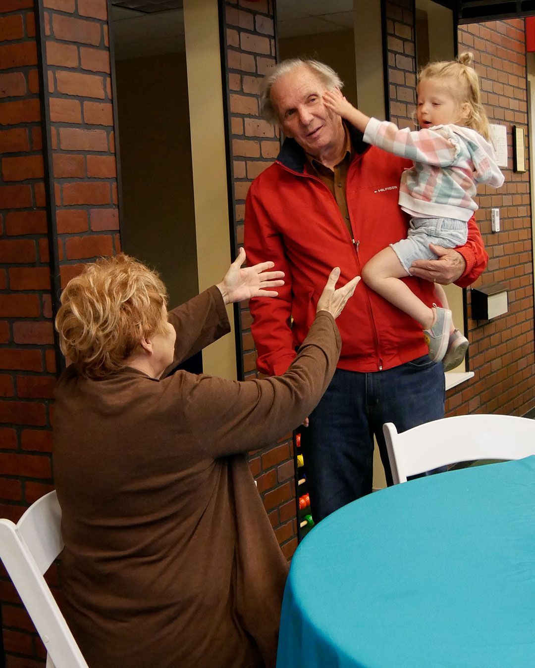 A grandfather holds his granddaughter while her grandmother reaches out to take her.