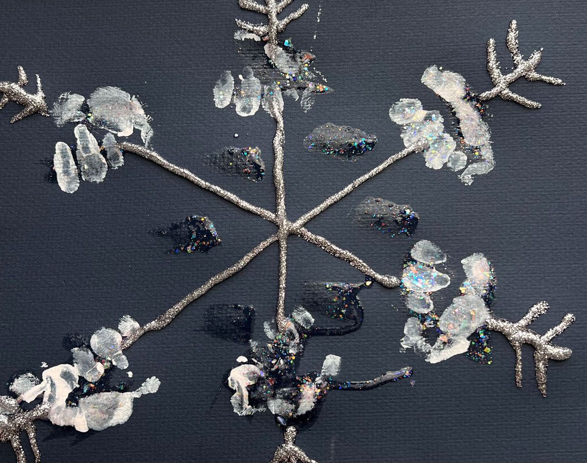 A six point child-made snowflake created with glitter glue and inward-pointing chevrons on the ends on black construction paper. The spines are decorated with handprints in white ink.
