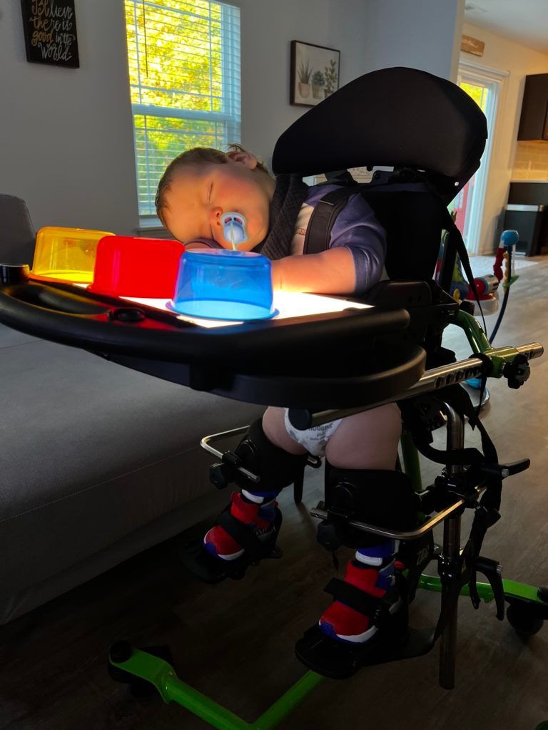 Lucas in his stander with a tray in front of him with light up toys. He is asleep with his head on the tray.