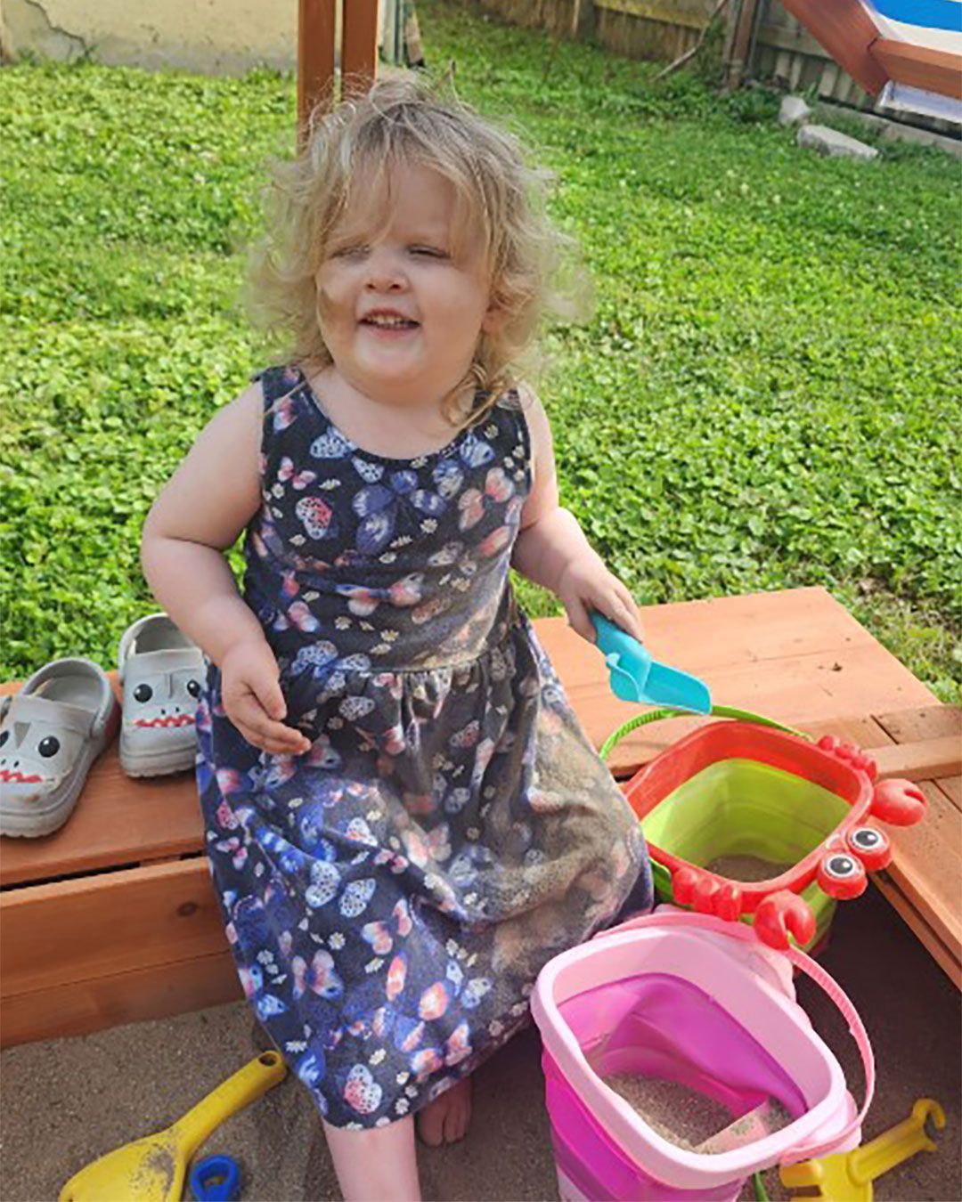 Roxanne smiles for the camera while sitting on the side of a sandbox and holding a shovel. Her feet are in the sand and there are pales and shovels.