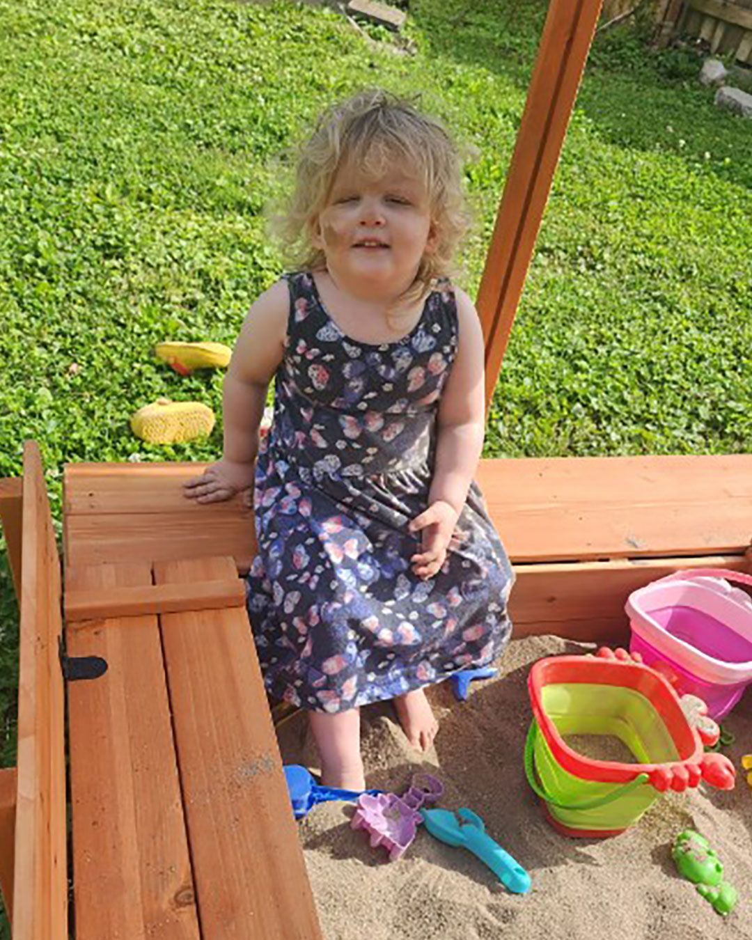 Roxanne smiles for the camera while sitting on the side of a sandbox. Her feet are in the sand and there are pales and shovels.