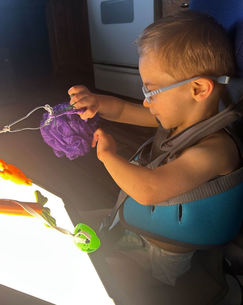 Elliot is smiling while wearing his glasses and pulling on objects attached to his light box.