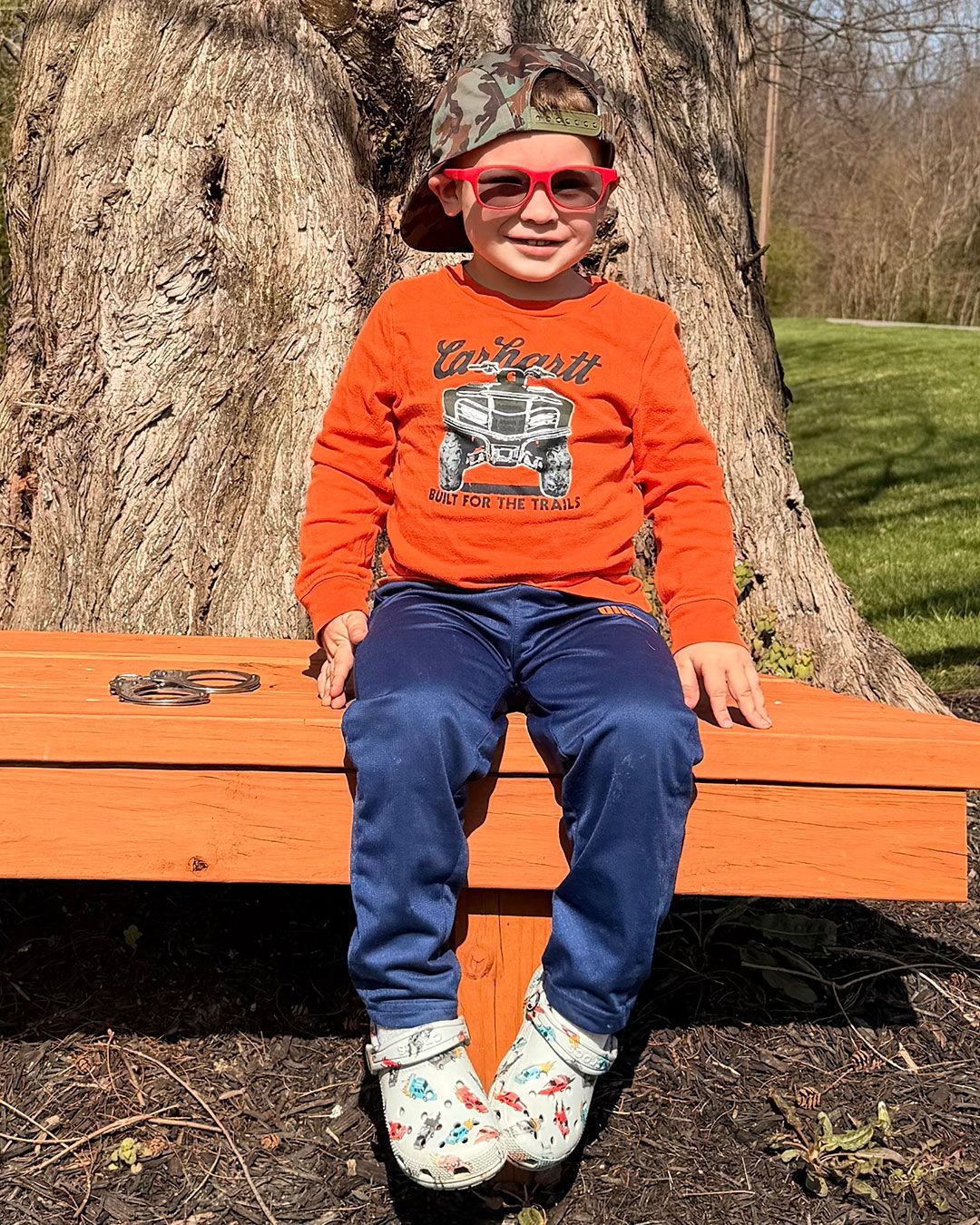 Wilder, wearing red prescription sunglasses, is sitting on a bench in the park and smiling for the camera.
