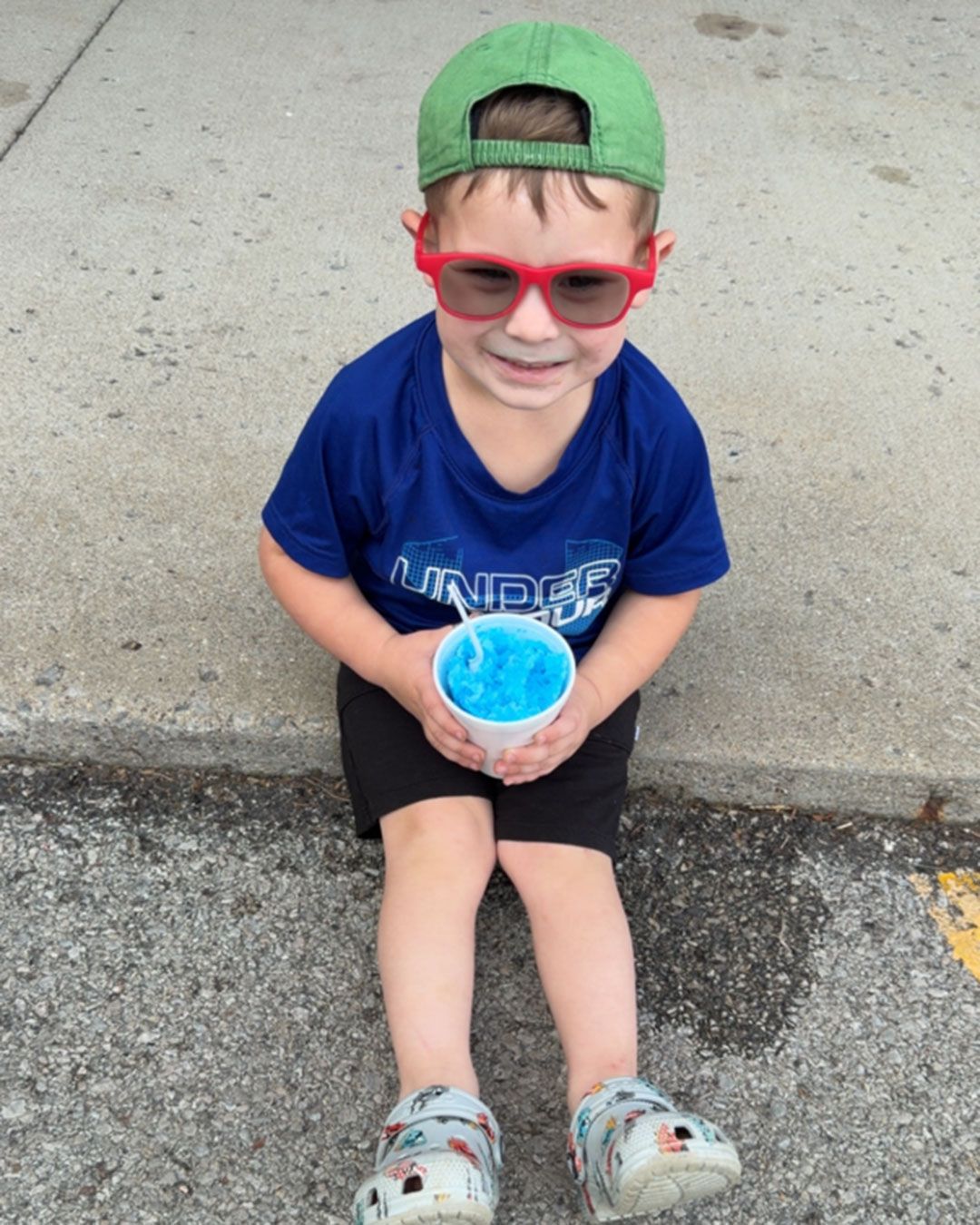 Wilder, wearing red prescription sunglasses, is sitting on the sidewalk holding a blue slushie and smiling for the camera.