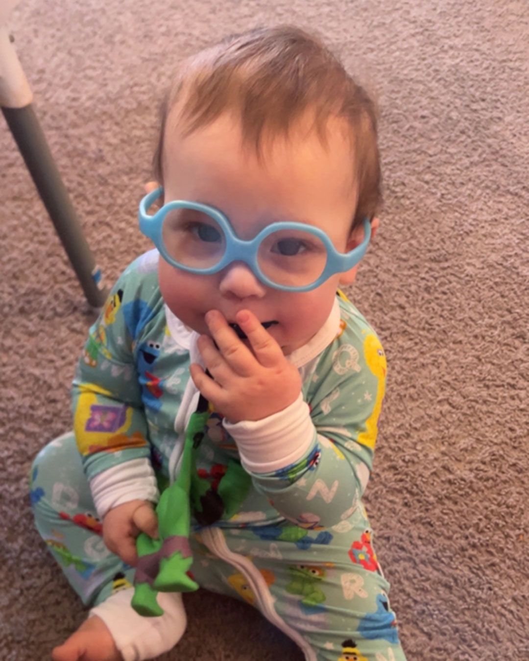 Wilder, wearing light blue glasses and his Sesame Street pjs, sits on the floor and smiles at the camera.