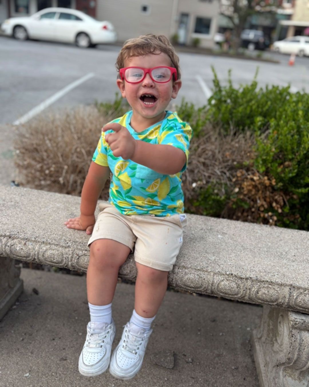 Wilder, wearing red glasses, is laughing and sitting on a concrete bench on a sidewalk.