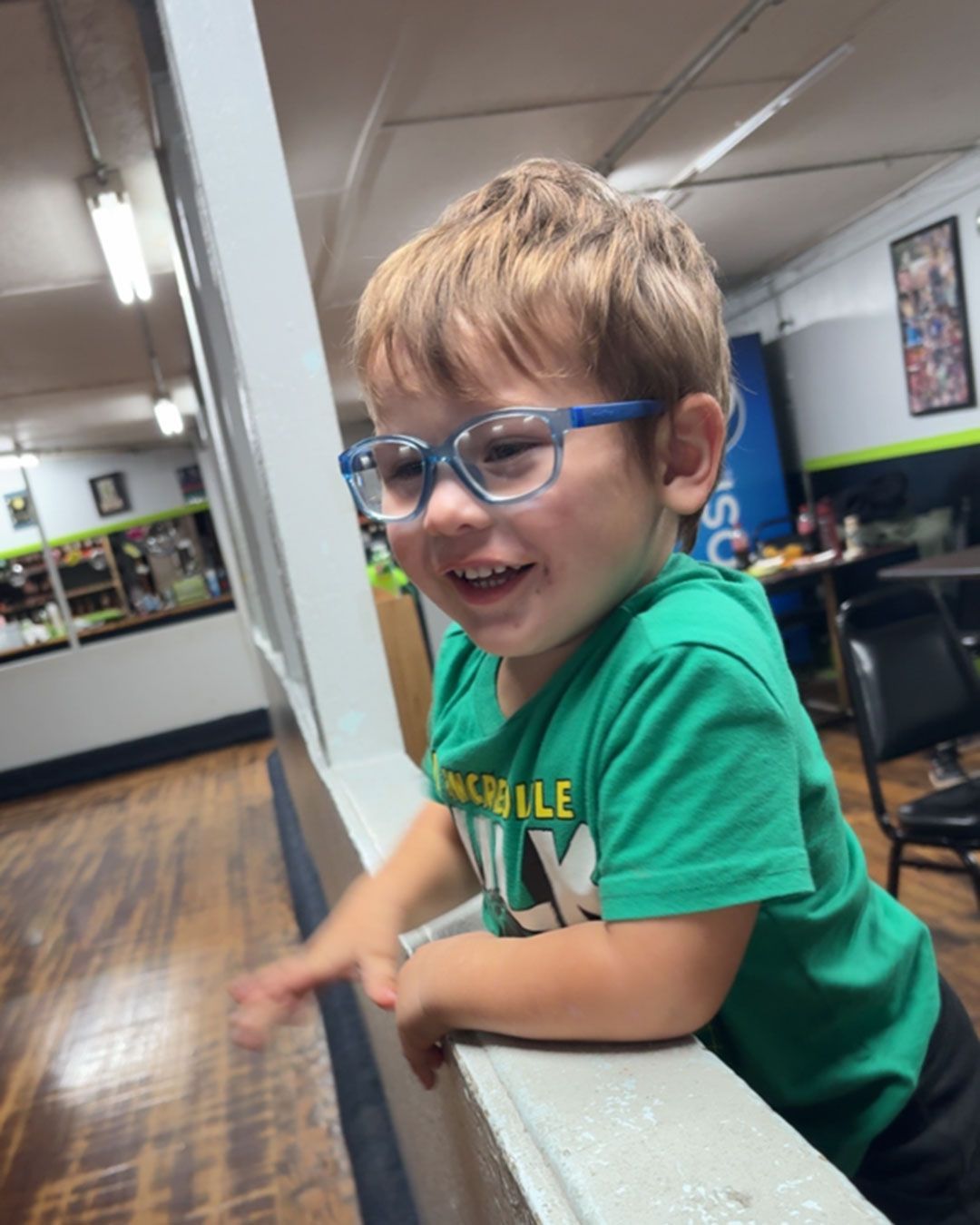 Wilder, wearing blue glasses, is laughing and leaning over a low wall in a restaurant.