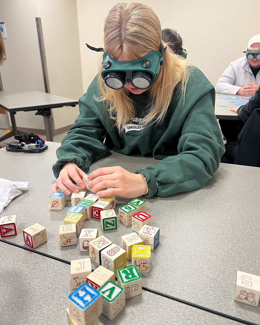 A female OT student sits at a table, holding alphabet blocks while wearing simulation goggles.