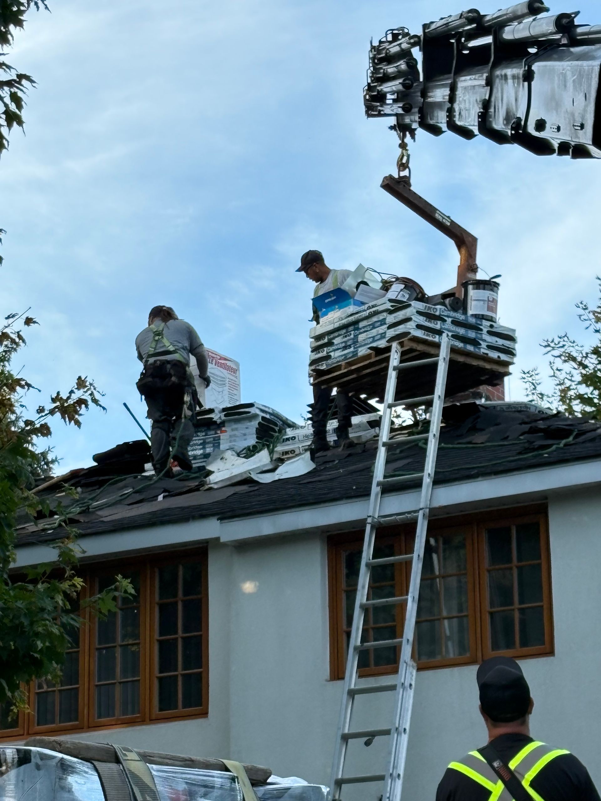 Des ouvriers sur un toit, à côté d'une échelle, manœuvrent une grue pour soulever des matériaux de couverture sur une maison.