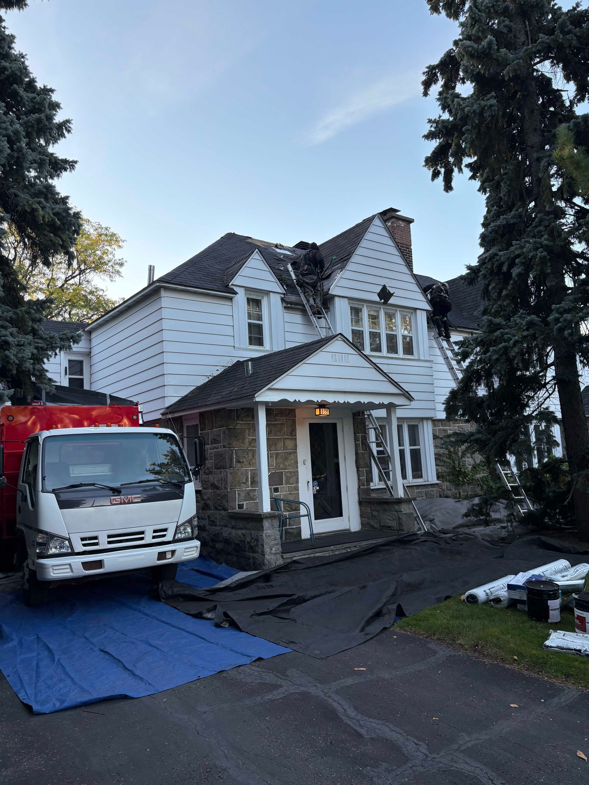 Maison blanche à deux étages avec toit foncé ; camion blanc garé dans l'allée à côté d'une bâche bleue et de grands arbres