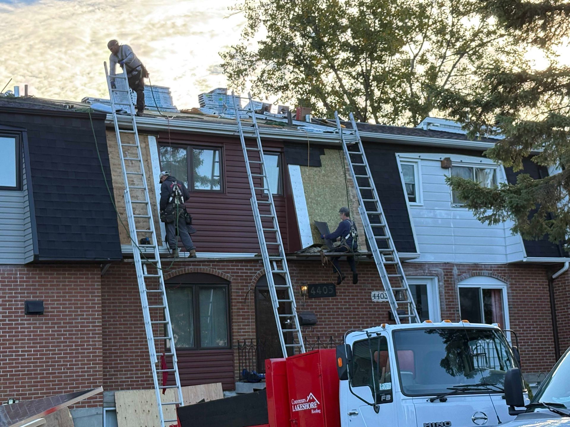 Des ouvriers, juchés sur des échelles, réparent le toit d'une rangée de maisons de ville en briques, un camion étant garé en dessous.