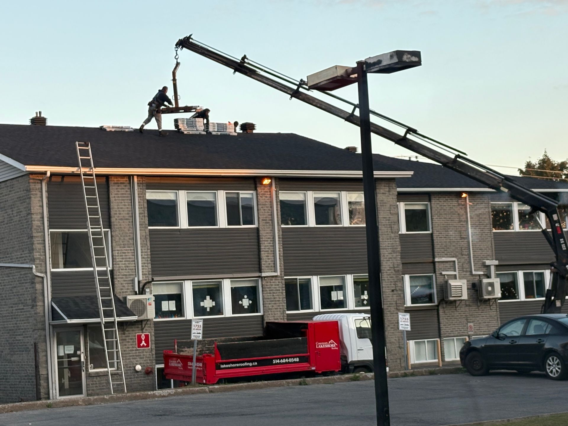 Des ouvriers sur le toit d'un immeuble d'appartements gris, une grue soulevant des matériaux au crépuscule.