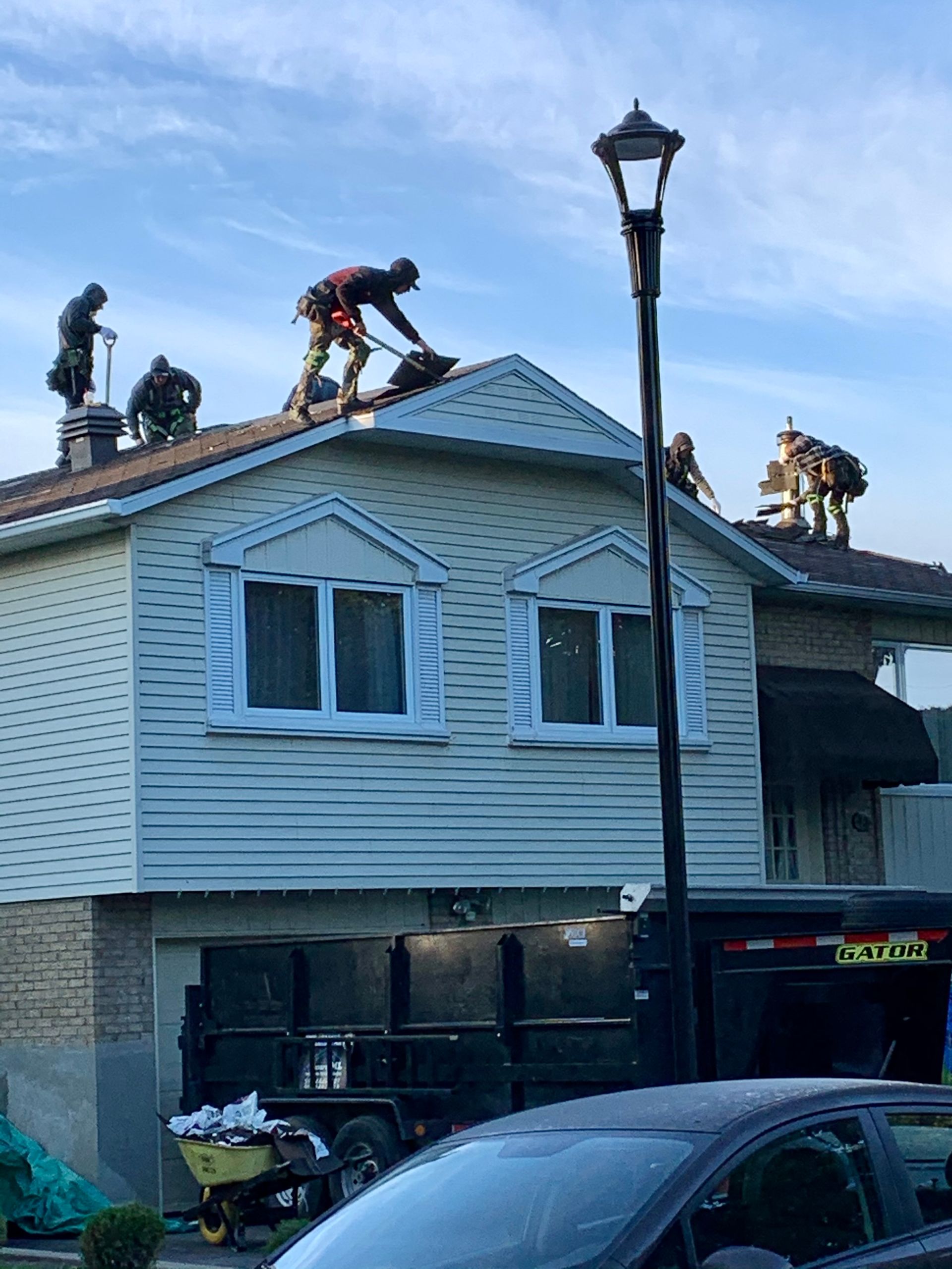 Des ouvriers réparent le toit d'une maison blanche, avec un lampadaire et des voitures devant.