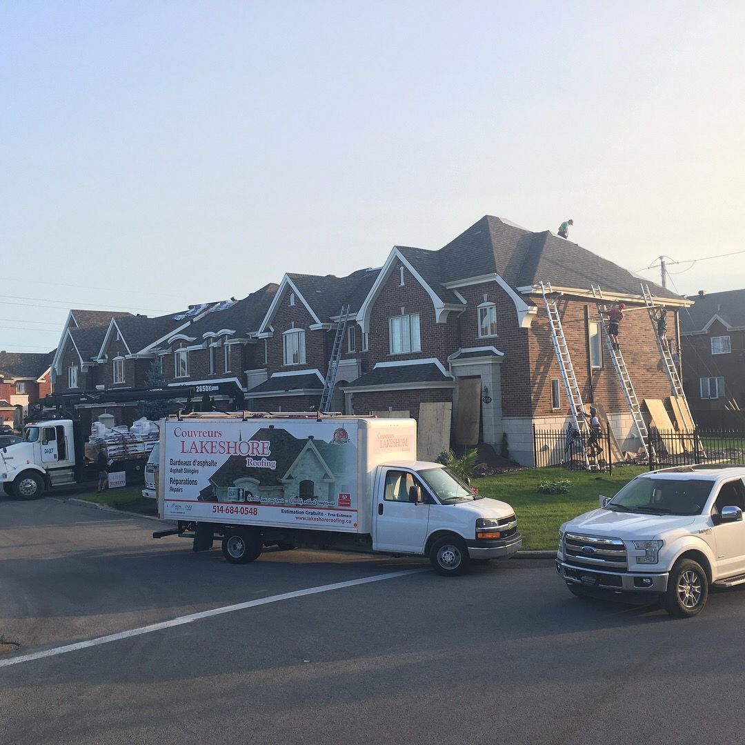 Rue résidentielle avec des fourgonnettes de chantier garées, une camionnette blanche et une rangée de maisons en briques en plein jour.