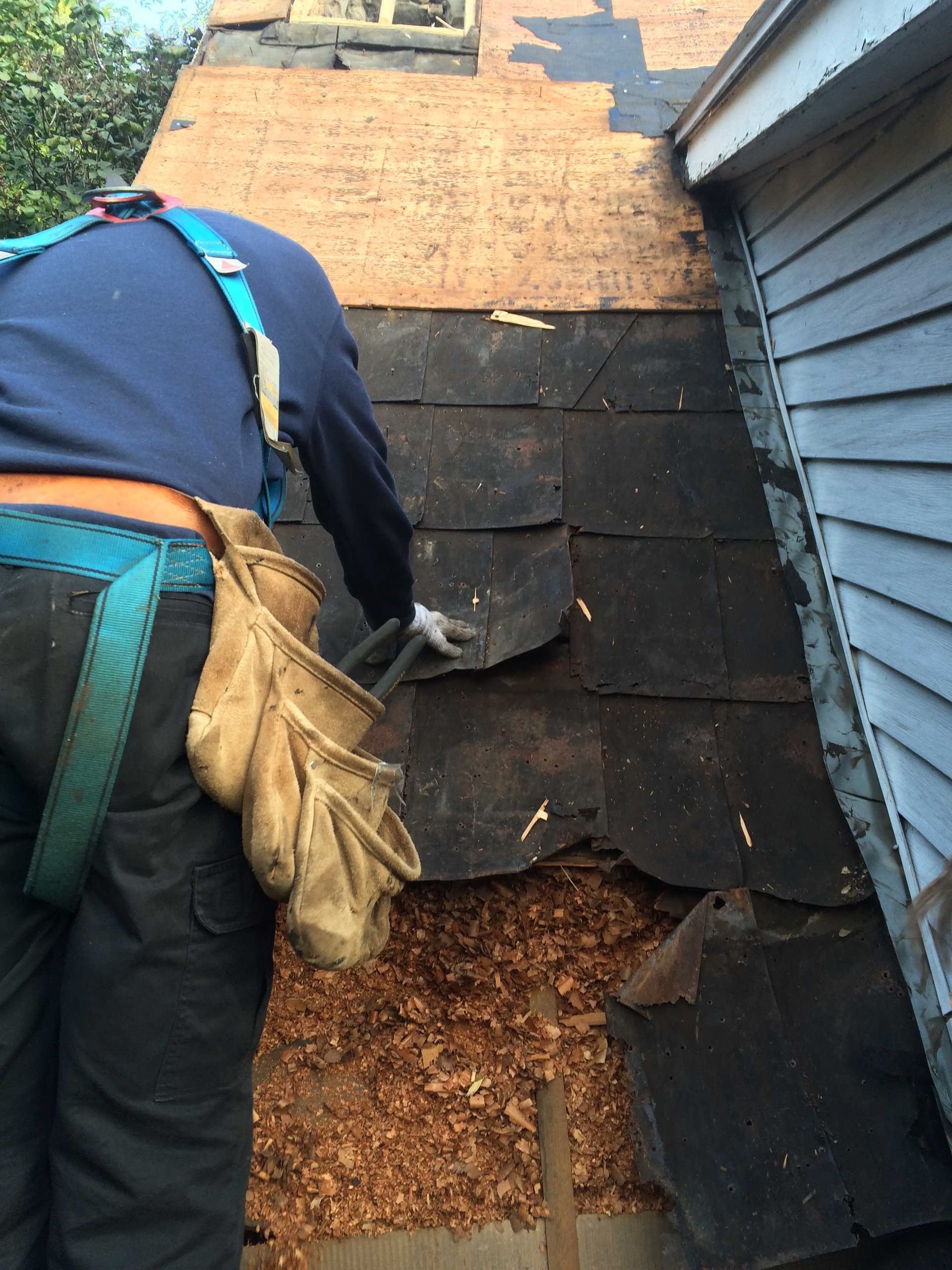 Un ouvrier répare des tuiles endommagées sur le toit d'une maison aux murs gris.