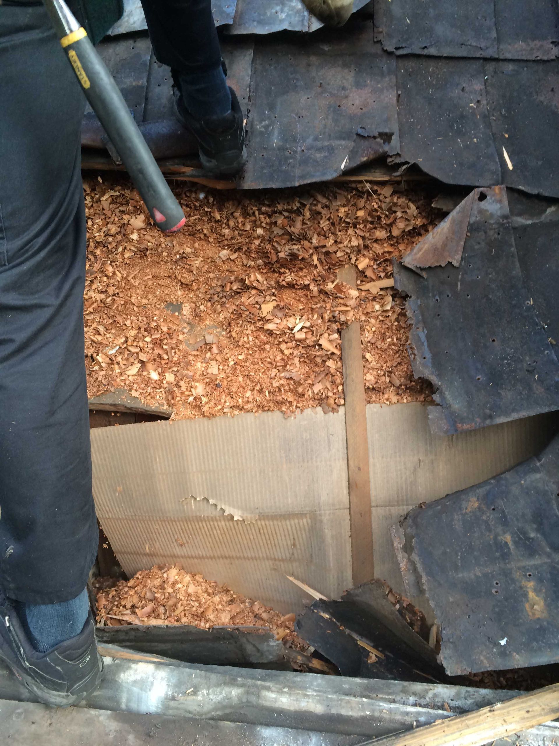 Toiture dont les bardeaux endommagés laissent apparaître du carton et des bardeaux de bois ; des bottes de travailleurs sont visibles à proximité.