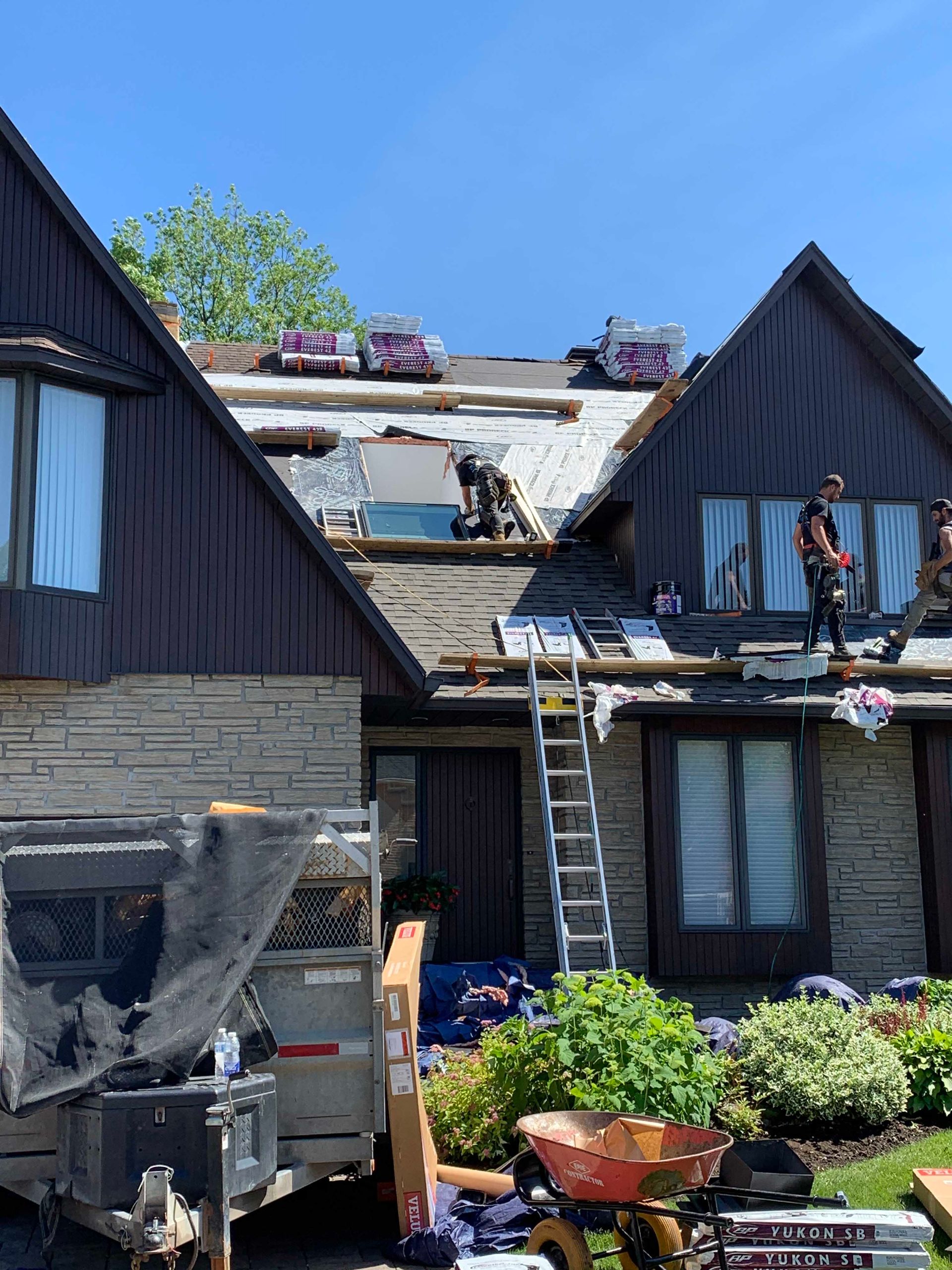 Des ouvriers travaillent sur un toit jouxtant des maisons en bois sombre, avec des échelles, des pots de fleurs et un camion en contrebas.